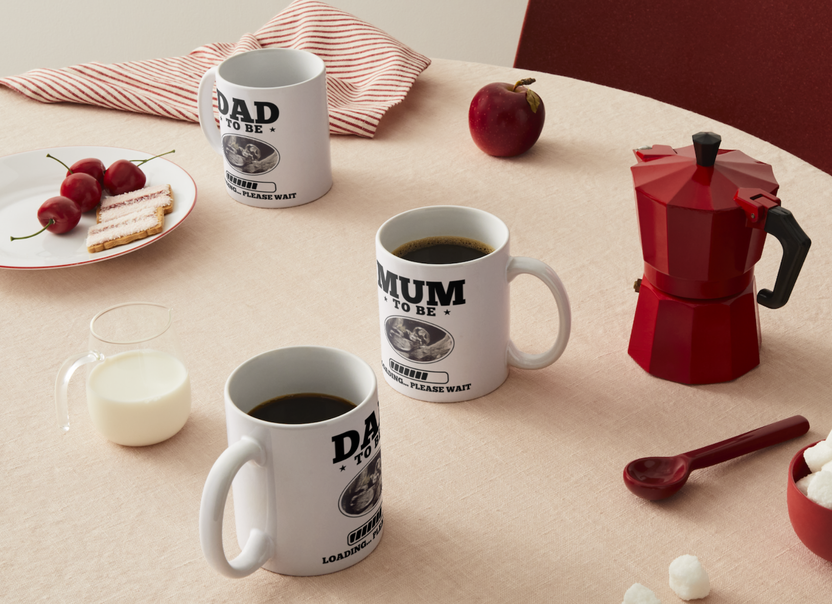 Three white mugs with 'DAD TO BE', 'MUM TO BE', and 'GRANDAD TO BE' text on a table with coffee and a red espresso maker.
