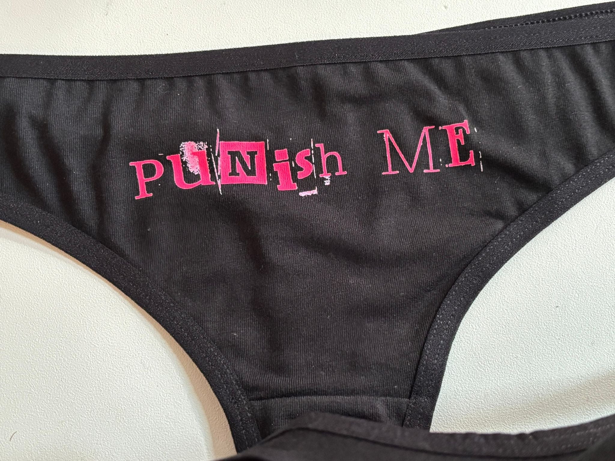 A close-up of a black sports bra featuring the word 'PUNISH' in distressed pink lettering on the front, laid flat on a light-colored surface.
