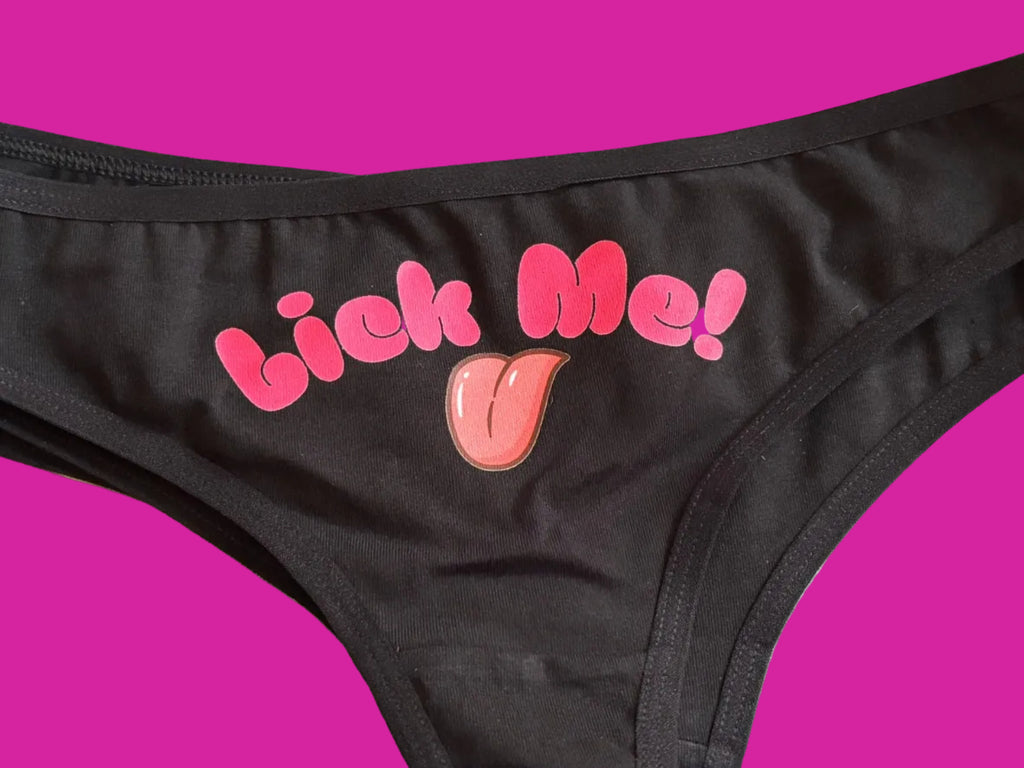 Close-up of a black bra or lingerie top with pink bubble lettering that reads 'lick me!' and a small pink cartoon tongue graphic on the front, laid flat on a light surface.