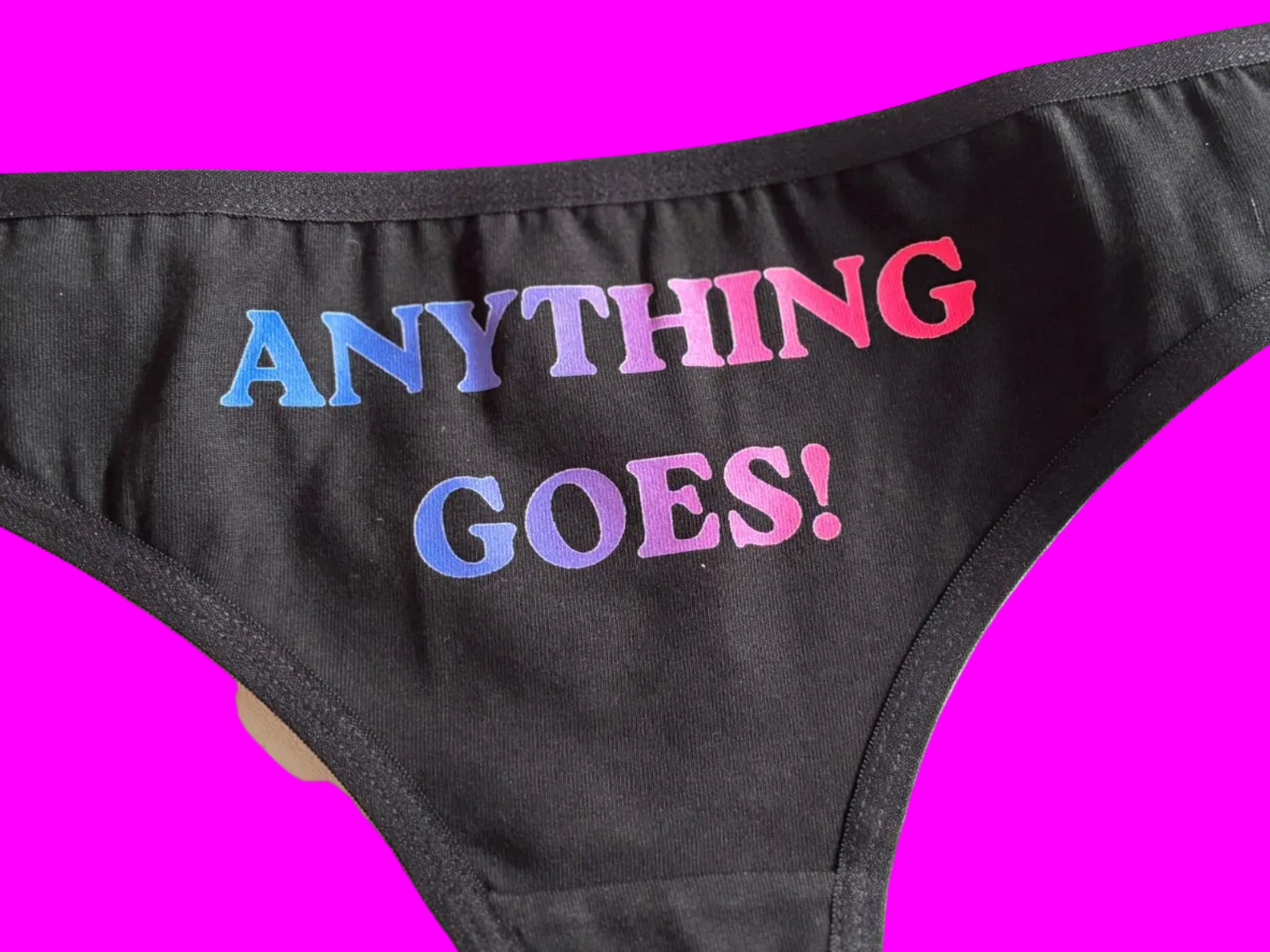 Black bikini bottoms featuring the phrase 'ANYTHING GOES!' printed vertically in a gradient of blue, purple, and pink, shown on a light-colored background.