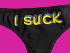 Close-up of black underwear with the word 'I SUCK' printed vertically in bold yellow letters outlined in pink and blue, on a light beige background.