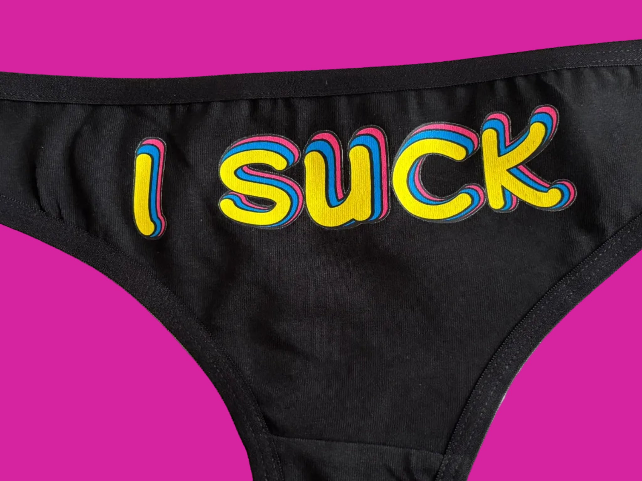 Close-up of black underwear with the word 'I SUCK' printed vertically in bold yellow letters outlined in pink and blue, on a light beige background.