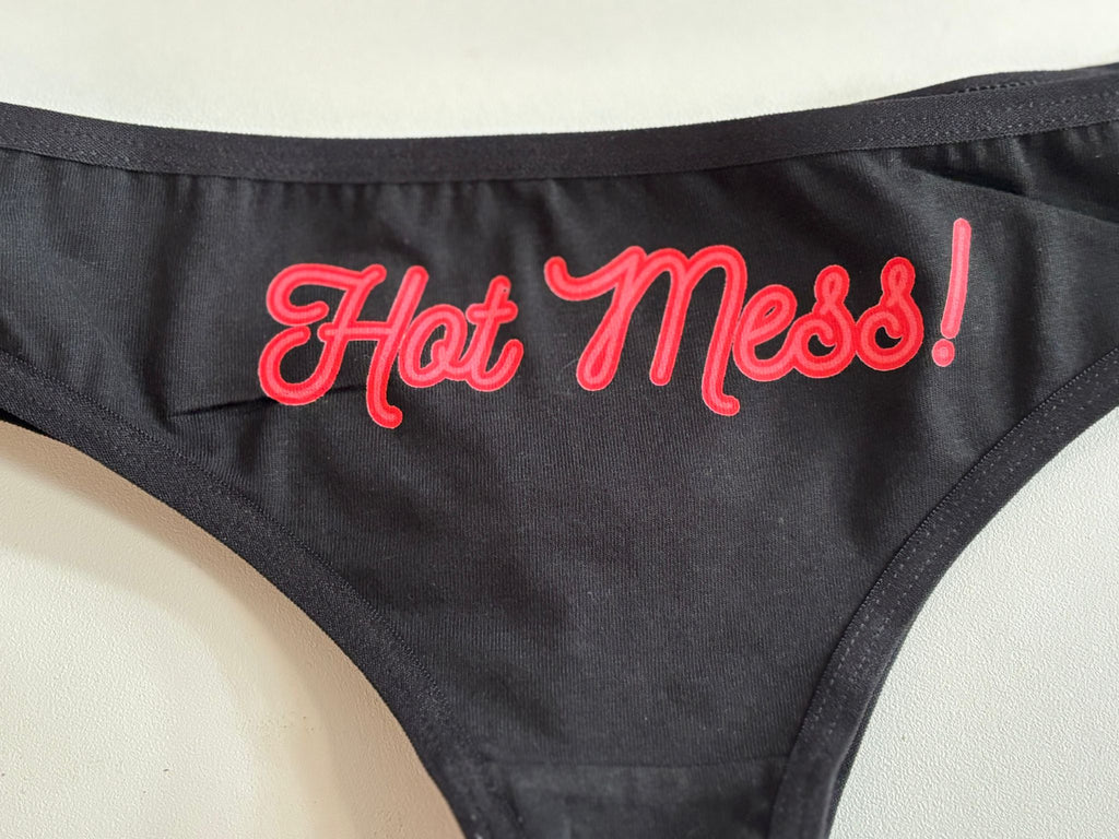 Close-up of a black bikini or swimwear bottom with bold red cursive text reading 'Hot Mess!' on the front panel, laid flat on a light surface.