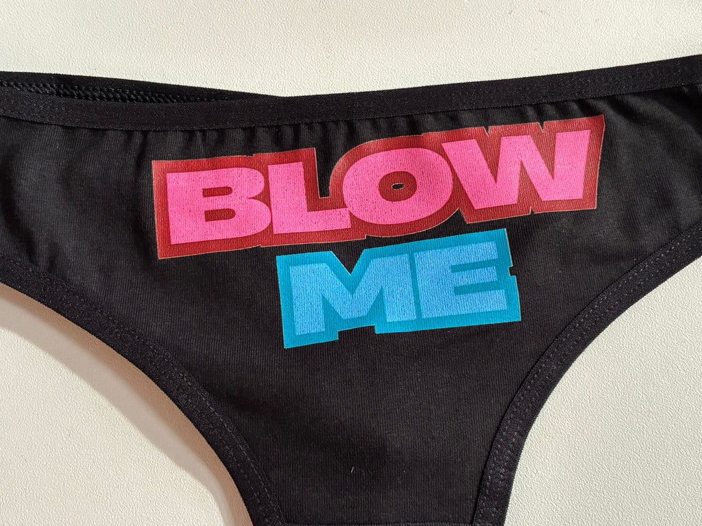 Close-up of a black garment featuring a large graphic text print that reads 'BLOW ME' in bold pink and blue lettering with red drop shadows, laid flat on a light beige surface.