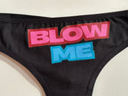 Close-up of a black garment featuring a large graphic text print that reads 'BLOW ME' in bold pink and blue lettering with red drop shadows, laid flat on a light beige surface.