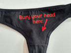 Black thong underwear with red text reading 'Bury your head here' and a red arrow pointing downward, shown on a light neutral background.