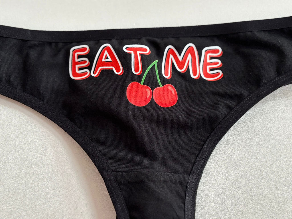 Black bikini bottom featuring a graphic print of two red cherries with green stems and the word 'EAT ME' in red letters with white outlines.