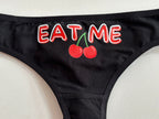 Black bikini bottom featuring a graphic print of two red cherries with green stems and the word 'EAT ME' in red letters with white outlines.