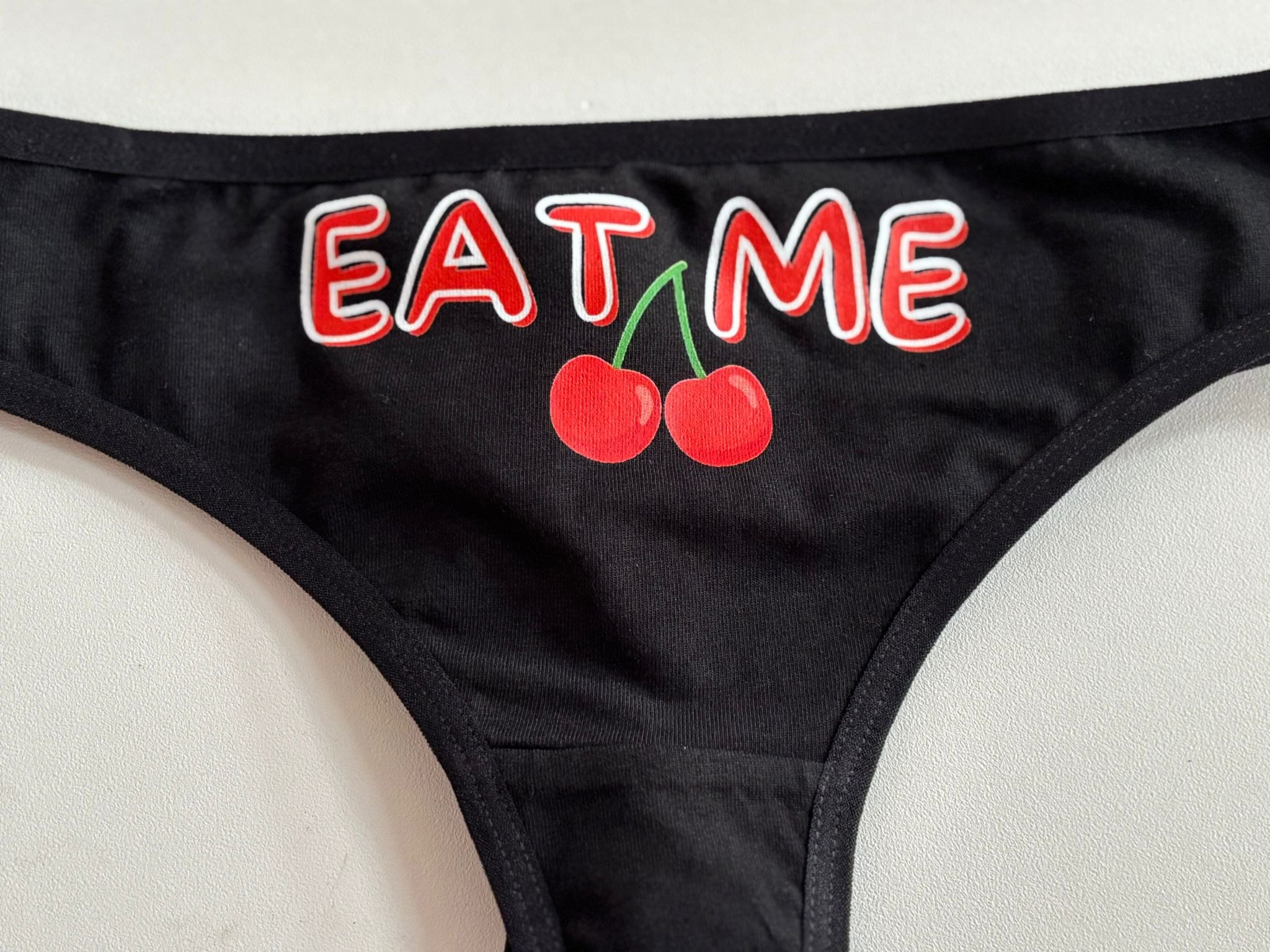 Black bikini bottom featuring a graphic print of two red cherries with green stems and the word 'EAT ME' in red letters with white outlines.