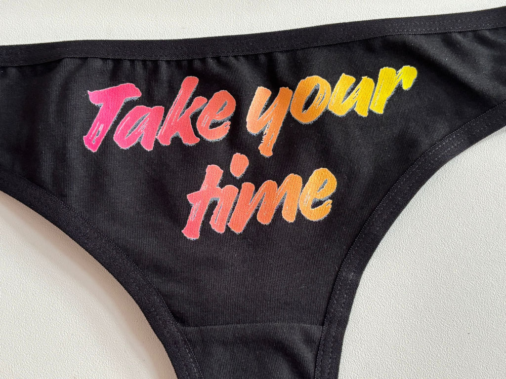 Close-up of a black bikini bottom with the phrase 'Take your time' printed in a colorful gradient font on the side.