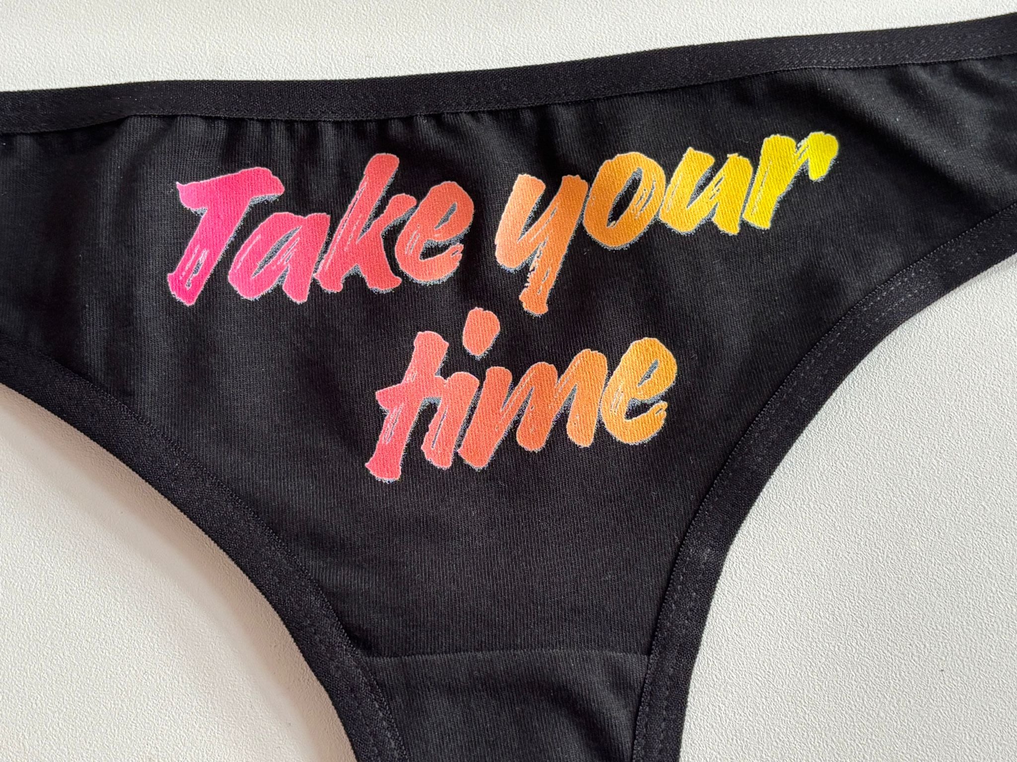 Close-up of a black bikini bottom with the phrase 'Take your time' printed in a colorful gradient font on the side.