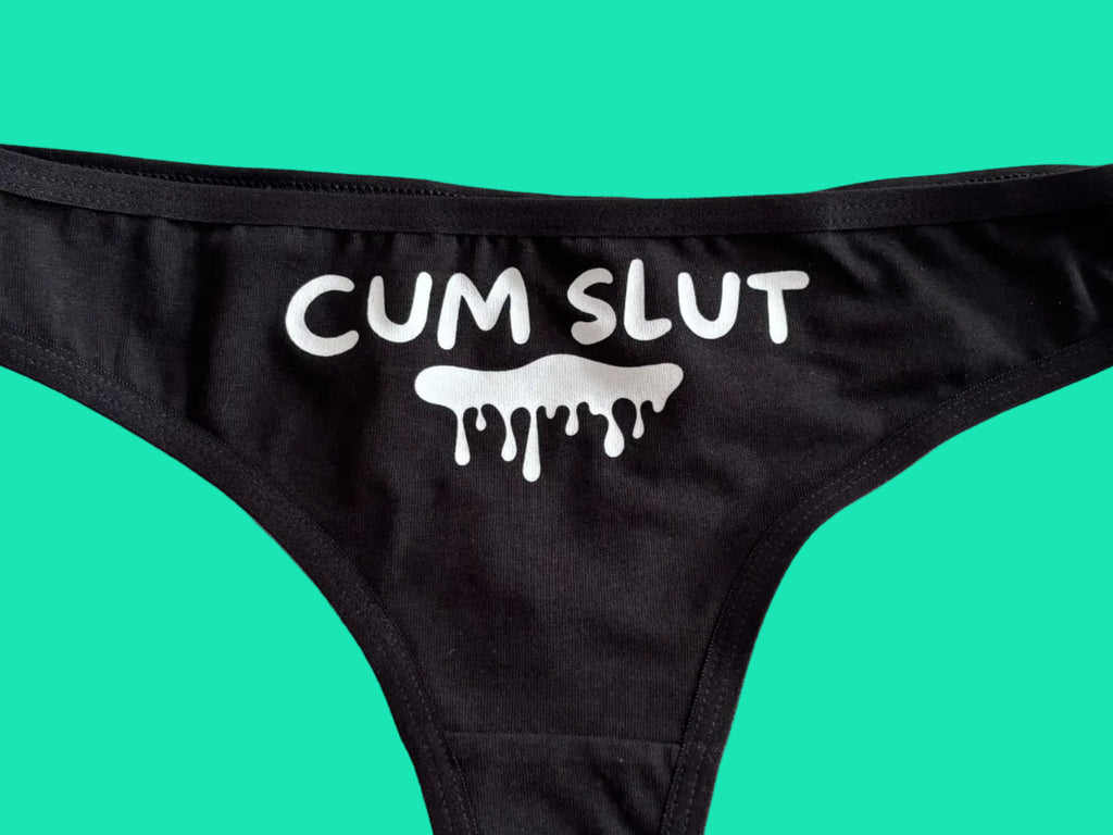 Black bikini bottom with a white graphic print reading 'CUM SLUT' and a dripping slime icon, laid flat on a light beige surface.