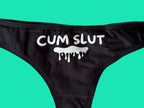 Black bikini bottom with a white graphic print reading 'CUM SLUT' and a dripping slime icon, laid flat on a light beige surface.