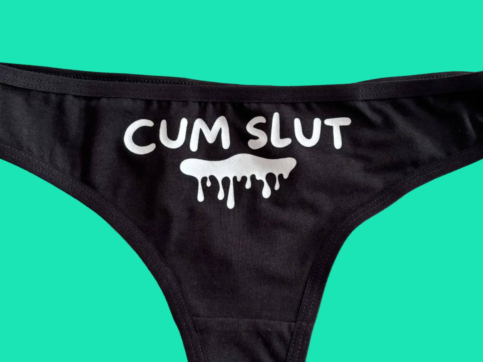 Black bikini bottom with a white graphic print reading 'CUM SLUT' and a dripping slime icon, laid flat on a light beige surface.