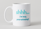 White mug with blue text 'shhh... I'm busy procrastinating!' on a light gray background